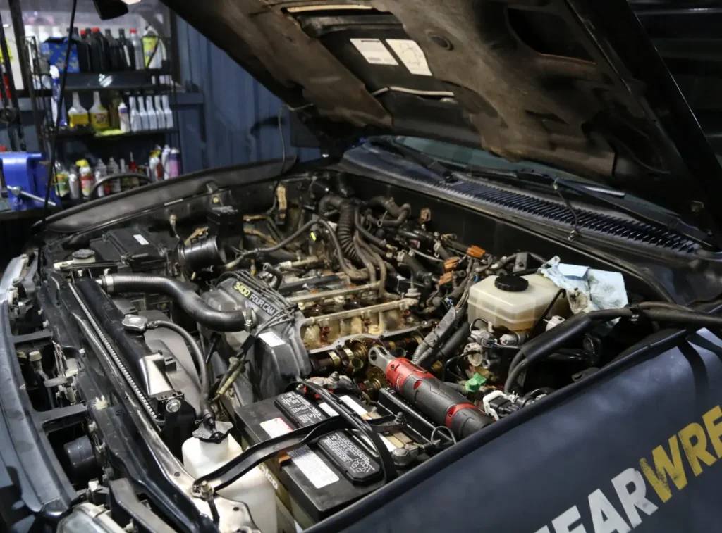 Japanese Motor Works: The open hood reveals the inner workings of a domestic auto repair scene, showcasing pipes, wires, and fluid reservoirs. Nearby are car tools and supplies on a shelf with bottles. The vehicles front displays partial lettering, emphasizing a focus on domestic auto care essentials.