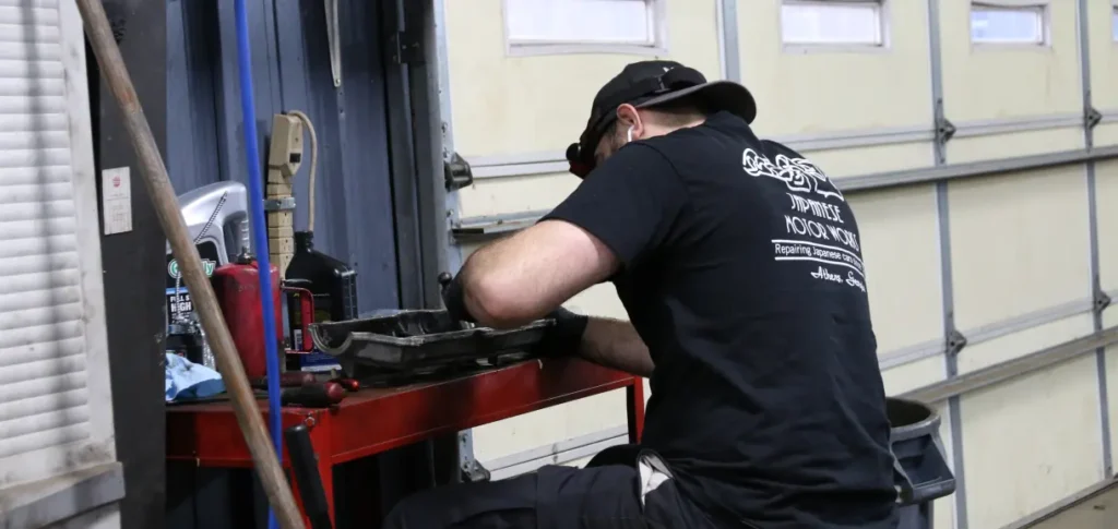 Japanese Motor Works: A person in a black cap and shirt works intently at a red workstation, handling a mechanical component for Honda repair in a garage. Tools, bottles, and a cleanup towel are nearby, with the garage doors visible in the background.